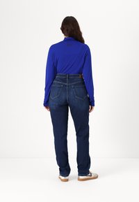 Camisa de manga longa azul-royal combinada com jeans escuros de cintura alta. Os jeans apresentam dois bolsos traseiros e uma etiqueta de pele castanha na cintura.