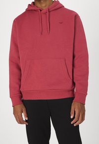 Felpa rossa in cotone con coulisse, tasca a marsupio, polsini e orlo a costine. Logo discreto sul petto. Indossata con pantaloni neri.