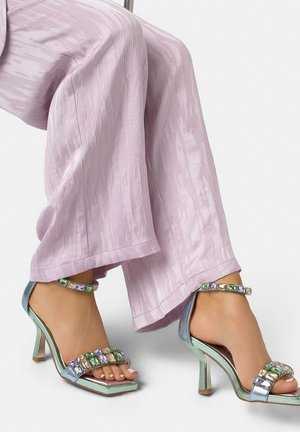 Pieds portant des sandales à talons hauts métalliques vertes avec des lanières ornées de pierres multicolores, assorties à un pantalon large texturé rose clair.