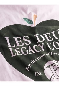 White cotton t-shirt with a green apple graphic, white text reading "LES DEUX LEGACY CO." and a small leaf detail.