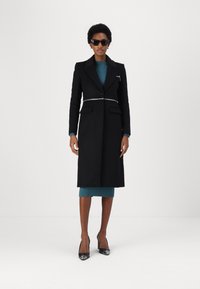 Cappotto di lana nera con un design strutturato, caratterizzato da un accento argento in vita, abbinato a un vestito a coste color teal e tacchi neri.