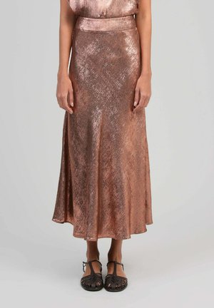 Femme portant une jupe midi brillante en or rose et des sandales plates noires tissées, debout devant un fond uni.