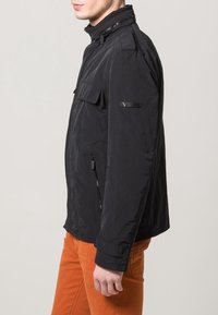 Czarna lekka kurtka z wysokim kołnierzem, kieszeniami na suwak i gładką teksturą; posiada naszywaną kieszeń na piersi oraz detal z logo.
