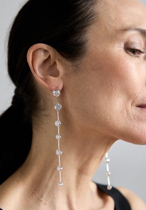 Ασημένια κρεμαστά σκουλαρίκια με στρογγυλους, διάφανους κρύσταλλους, που διαθέτουν γραμμικό σχέδιο και εναλλασσόμενη τοποθέτηση κρυστάλλων κατά μήκος μιας λεπτής αλυσίδας.