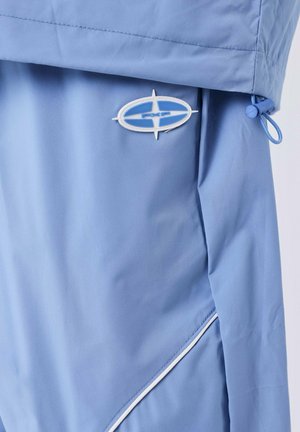 Chaqueta azul claro con un logotipo ovalado PXP en blanco y azul, un cordón con ajuste y detalles de piping blanco en la costura lateral.