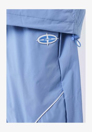 Veste bleu clair avec un logo ovale PXP blanc et bleu, un cordon de serrage avec un loquet, et un détail de piping blanc sur la couture latérale.