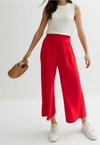 Red wide-leg pants with a high waist, paired with a textured white sleeveless top and white sneakers; holding a woven bag.