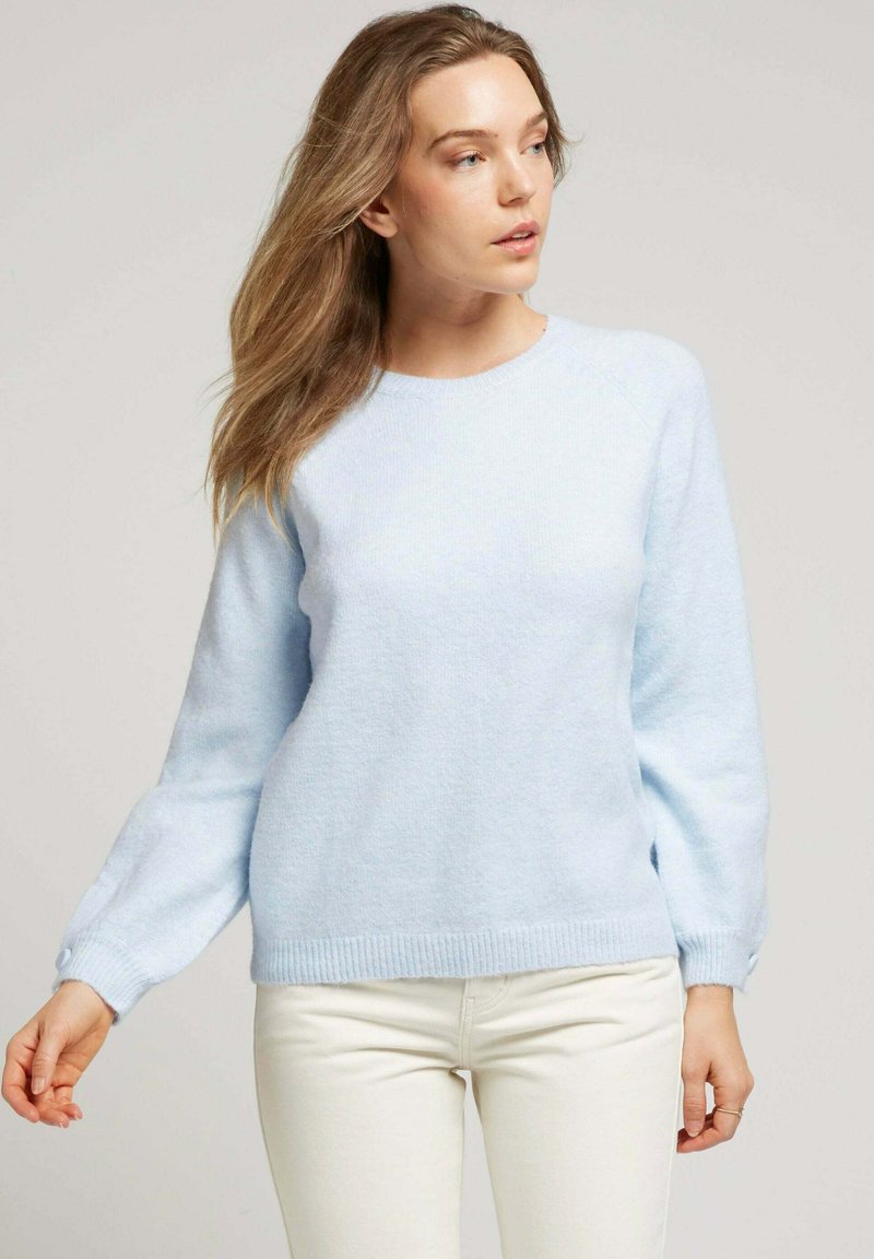 Pull en tricot doux bleu clair avec un encolure ronde et de longues manches légèrement bouffantes. Détails à bords côtelés au niveau de l'ourlet et des poignets. Porté avec un pantalon blanc.