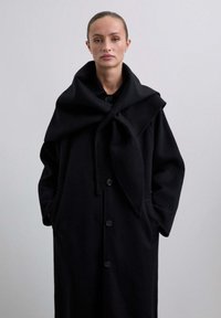 Abrigo largo negro de tela suave con un cuello grande y drapeado, botones frontales y bolsillos laterales. Textura lisa y diseño minimalista.