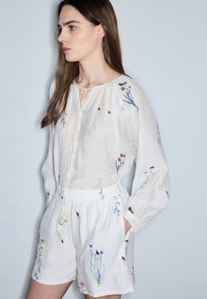 Mujer que lleva una blusa blanca de manga larga y pantalones cortos con delicados bordados florales multicolores, de pie con una mano en el bolsillo, mirando hacia la izquierda.
