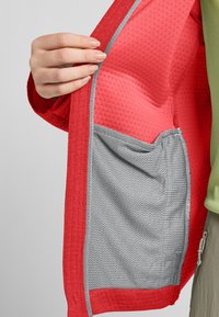Rote Jacke mit einer strukturierten Außenseite und glatter Innenseite, ausgestattet mit einer grauen Mesh-Tasche. Futter ist leuchtend rot mit einem gepunkteten Muster.