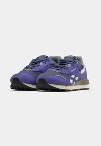 Pāris purpura un pelēku Reebok sporta apavu ar baltām svītrām un melnā zolē, vērsti uz priekšu uz baltā fona.