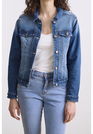 Frau trägt eine hellblaue Jeansjacke über einem weißen Tanktop, kombiniert mit hellblauen Jeans, vor einfachem Hintergrund.