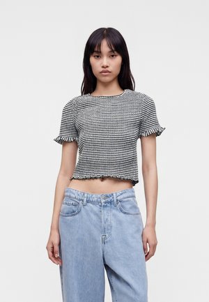 Junge Frau trägt ein schwarz-weiß gestreiftes kurzärmeliges Croptop mit Rüschensaum und hellblaue High-Waist-Jeans vor einfarbigem Hintergrund.