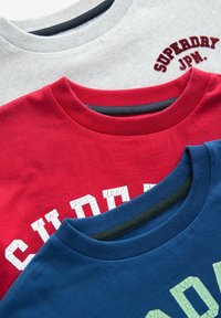 Tres camisetas de cuello redondo apiladas: gris con bordado, roja con letras blancas agrietadas y azul marino con texto verde claro. Tela de algodón.