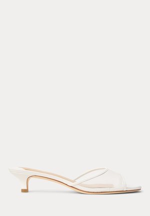 Sandalia mule blanca de tacón bajo con una tira transparente y diseño de punta abierta sobre un fondo liso.