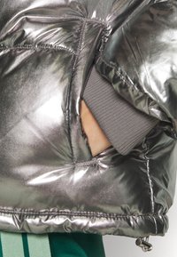 Stříbrná metalická péřová bunda s prošívanou texturou, vybavená nenápadnou boční kapsou a světle šedými žebrovanými manžetami pro větší pohodlí.