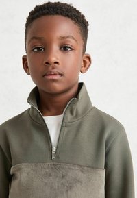 Sudadera de medio cierre en verde oliva con un panel en el pecho suave y parecido al ante. Presenta un cuello alto y una superficie texturizada.