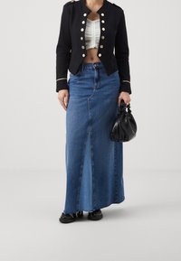 Black military-style jacket with gold buttons, white ruched top, blue denim maxi skirt, and black textured handbag. Flat black shoes.