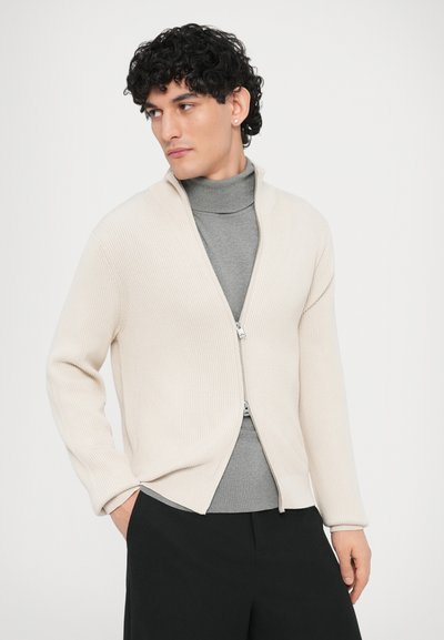 Κρεμ ζακάρ ζακέτα με φερμουάρ και μεγάλο γιακά, φορεμένη πάνω από γκρι ζιβάγκο. Το μοντέλο φοράει μαύρο παντελόνι, με τα χέρια στις τσέπες, κοιτάζοντας ελαφρώς πλάγια.