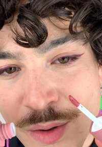 Flytande läppfärg i en rosa tube med en vit applicator. Personens ansikte har bruna ögon, en mustasch och lockigt mörkt hår.