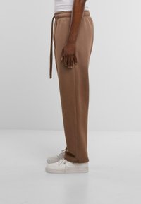 Braune Sweatpants mit elastischem Bund, Kordelzug-Detail und geradem Schnitt, kombiniert mit weißen Sneakern. Glatte Textur.
