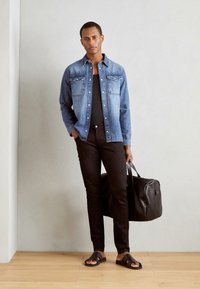 Kék farmerekből készült gombos ing, fekete póló, fekete slim-fit nadrág, fekete bőr duffel táska és fekete szandál, két pánttal.