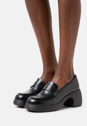 Mocassins en cuir noir avec un talon plateforme et un design courbé. Comprend une finition lisse et des détails cousus sur la tige.