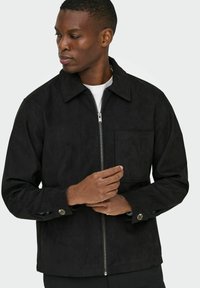 Črna suknjič iz semiša s ovratnikom, sprednjim zadrgo, žepom na prsih in gumbnimi manšetami. Gladka tekstura in sproščen kroj.