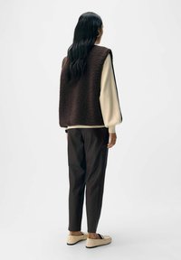 Gilet in pile marrone con vestibilità comoda, camicia a maniche lunghe crema, pantaloni attillati marrone scuro e mocassini di colore chiaro. Design e texture semplici.
