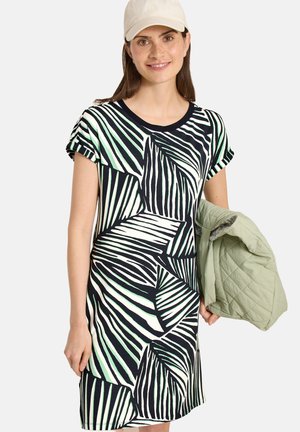 Femme portant une robe rayée noire et blanche et une casquette beige, tenant une veste matelassée vert clair dans sa main gauche.