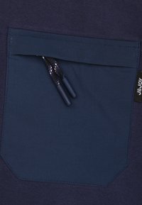 Marineblauwe stof met een zak met een contrasterende textuur, versterkte stiksels en details met een trekkoord met plastic uiteinden.