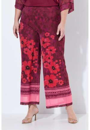 Spodnie o szerokich nogawkach z dużymi czerwonymi kwiatowymi wzorami na ciemnym bordowym materiale oraz różową geometryczną bordiurą, noszone z cielistymi sandałami na wysokim obcasie.