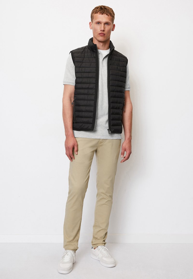 Gilet sans manches rembourré noir avec fermeture zippée, porté sur un polo gris clair et un pantalon beige, associé à des baskets blanches.