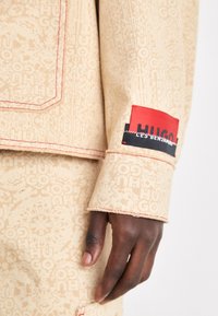 Beige tygjacka med en subtil mönstrad textur. Har röda sömmar och en kontrasterande röd och svart etikett på manschetten.