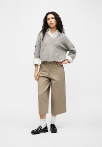 Γυναίκα με σγουρά μαλλιά στέκεται με το χέρι στη μέση, φοράει γκρι κοντό πουλόβερ πάνω από λευκό πουκάμισο, μπεζ παντελόνια με φαρδιά μπατζάκια, λευκές κάλτσες και μαύρα παπούτσια.