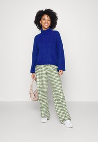 Maglione blu a collo alto lavorato a maglia e pantaloni verdi a motivo houndstooth. Il modello tiene una borsa beige; le sneakers bianche completano l'outfit.
