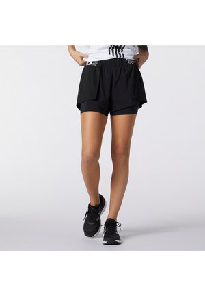 Osoba nosząca czarne, warstwowe sportowe spodenki i czarne buty do biegania, stojąca ze skrzyżowanymi nogami na jednolitym tle.