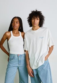 Débardeur blanc à côtes associé à un jean taille haute bleu clair ; homme en t-shirt blanc oversized avec texte bleu clair, affichant un style décontracté.