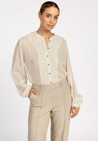 Crème blouse met knoopsluiting en een textuur aan de voorkant, geruite patronen op de mouwen en een relaxte pasvorm. Gecombineerd met beige op maat gemaakte broeken.