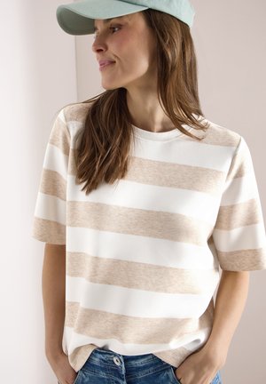 Femme aux longs cheveux bruns portant un t-shirt à manches courtes beige et blanc à rayures, une casquette bleu clair et un jean bleu, debout les mains dans les poches.