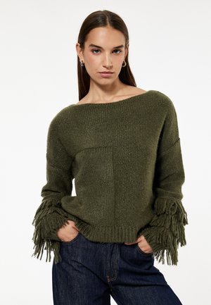 Kobieta ubrana w zielony sweter dziergany z frędzlami na rękawach i ciemnoniebieskie jeansy, stojąca z rękami w kieszeniach na białym tle.