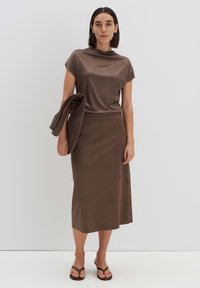 Bruin top met korte mouwen en gedrapeerde halslijn, in combinatie met een bruine kokerrok. Het model houdt een bijpassende textuurblazer vast. Platte sandalen maken de look compleet.