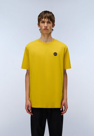 Gul bomulds t-shirt med korte ærmer, rund halsudskæring og et lille, cirkulært sort logo på brystet. Enkelt design, ingen mønstre.