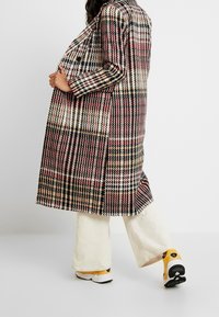 Károvaný kabát v krémové, burgundské a černé barvě s uvolněným střihem, dlouhými rukávy a délce ke kolenům. Spárován s krémovými manšestrovými kalhotami a teniskami.