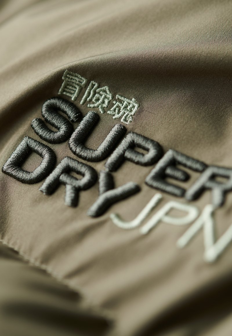 Gestickter Stoffaufnäher mit "SUPER DRY" in Dunkelgrau und "JPN" in Hellgrün auf einem strukturierten khakifarbenen Hintergrund.