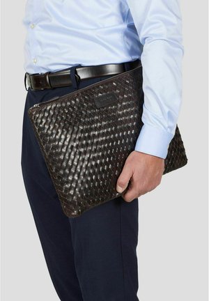 Mann i blå skjorte og mørke bukser som holder en stor, mørk brun flettet skinnpung med glidelås under armen.