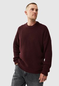 Burgundy knit sweater with a textured surface, wide crew neck, and ribbed cuffs, paired with gray jeans.
