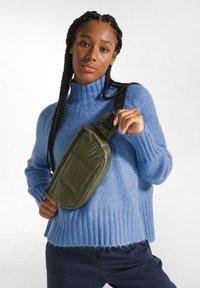 Borsa a tracolla verde in nylon con texture trapuntata, dotata di chiusura con zip e tracolla regolabile. Indossata sopra un maglione azzurro chiaro.