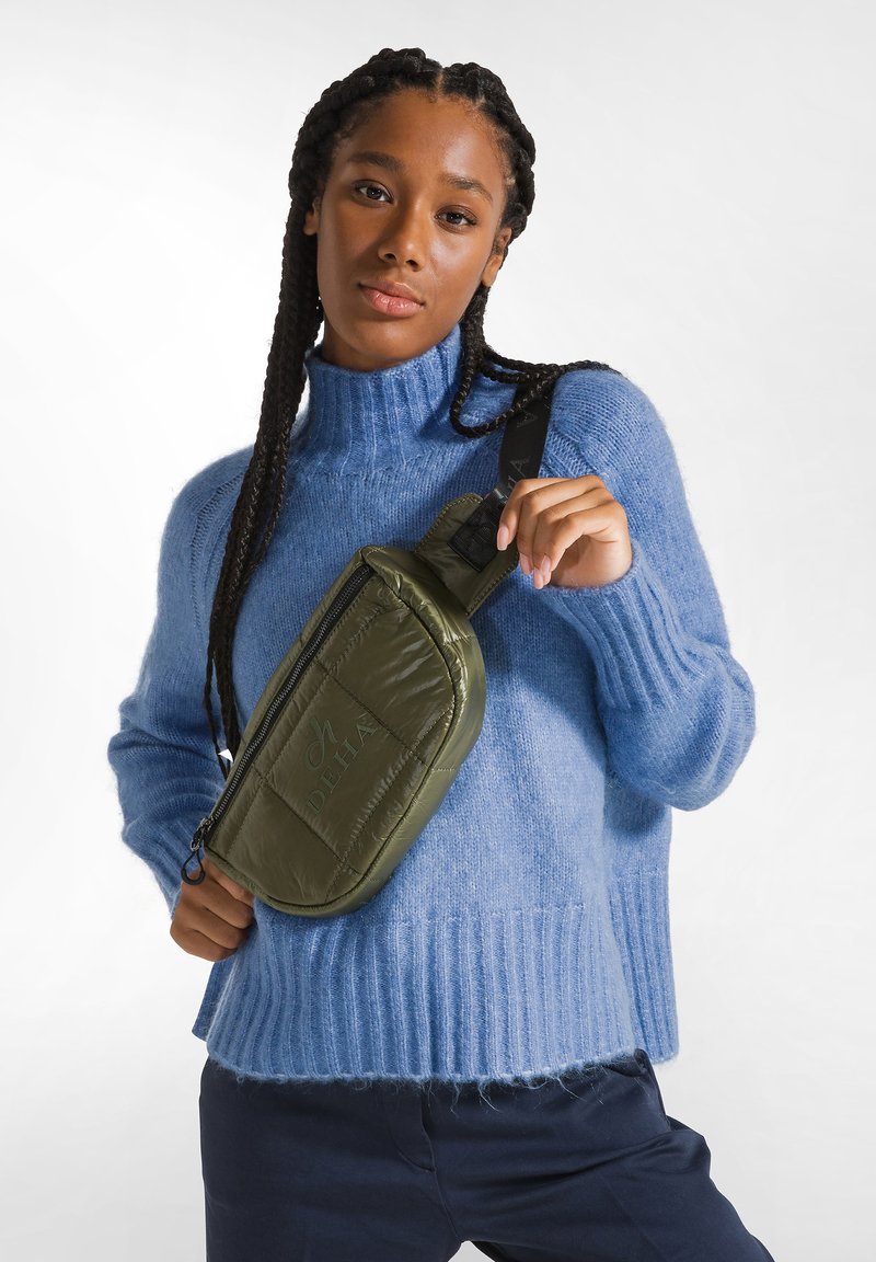 Borsa a tracolla verde in nylon con texture trapuntata, dotata di chiusura con zip e tracolla regolabile. Indossata sopra un maglione azzurro chiaro.